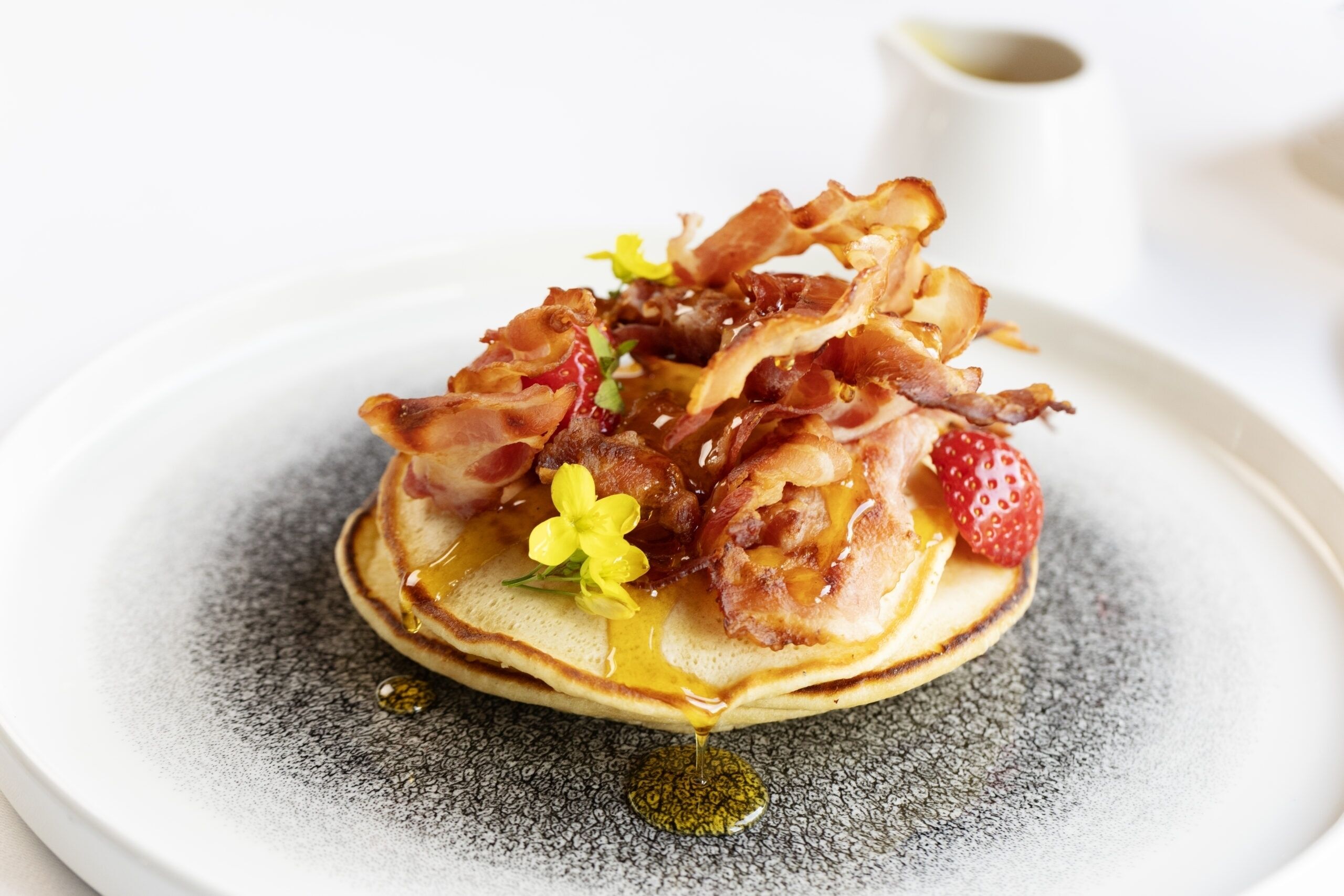 Una pila de panqueques cubiertos de tocino, fresas y una flor pequeña, servidos en un plato con textura.