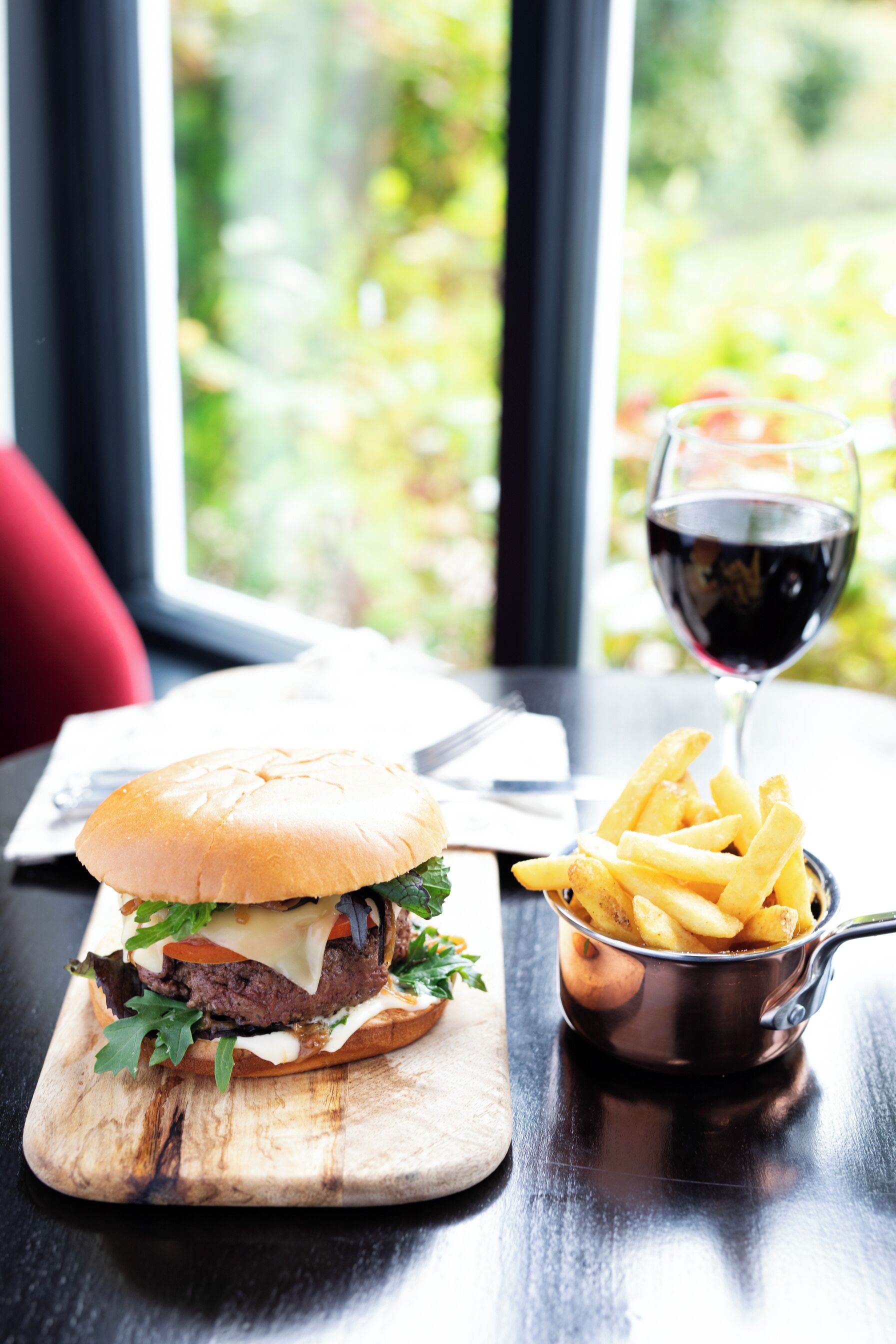 A meal with a burger and fries on a wooden board, a glass of red wine, and a window with greenery outside.