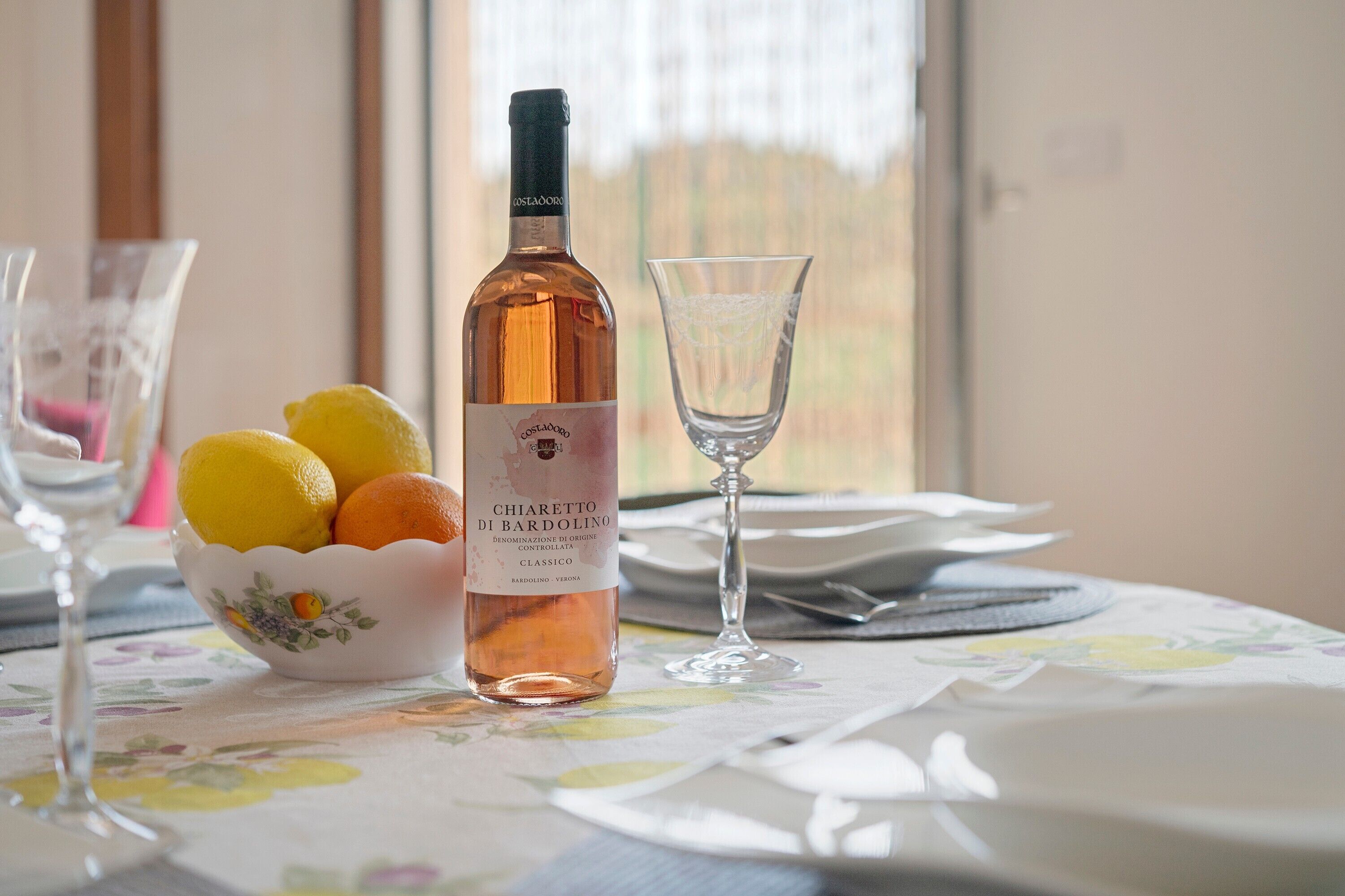 Une bouteille de vin Chiarolet di Bardolino, un bol de citrons et un verre à vin sur une table recouverte d’une nappe à motifs floraux.
