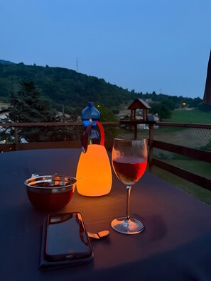 A table with a smartphone, a glass of red wine, a bowl of cherries, and a blue water bottle on a balcony overlooking a scenic landscape.