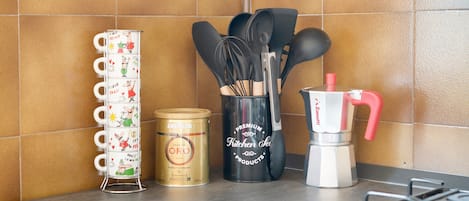 A kitchen counter with a coffee maker, a can of pasta, a set of whisks, and a mug rack.