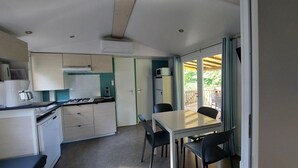 A compact kitchen with a gas stove, microwave, and a small refrigerator. There is a dining area with a table and chairs, and a sliding glass door leading to an outdoor deck. - Premium 2 Mobile Home with Mountain View, Wi-Fi, and Air Conditioning (Saint-Laurent-en-Beaumont)