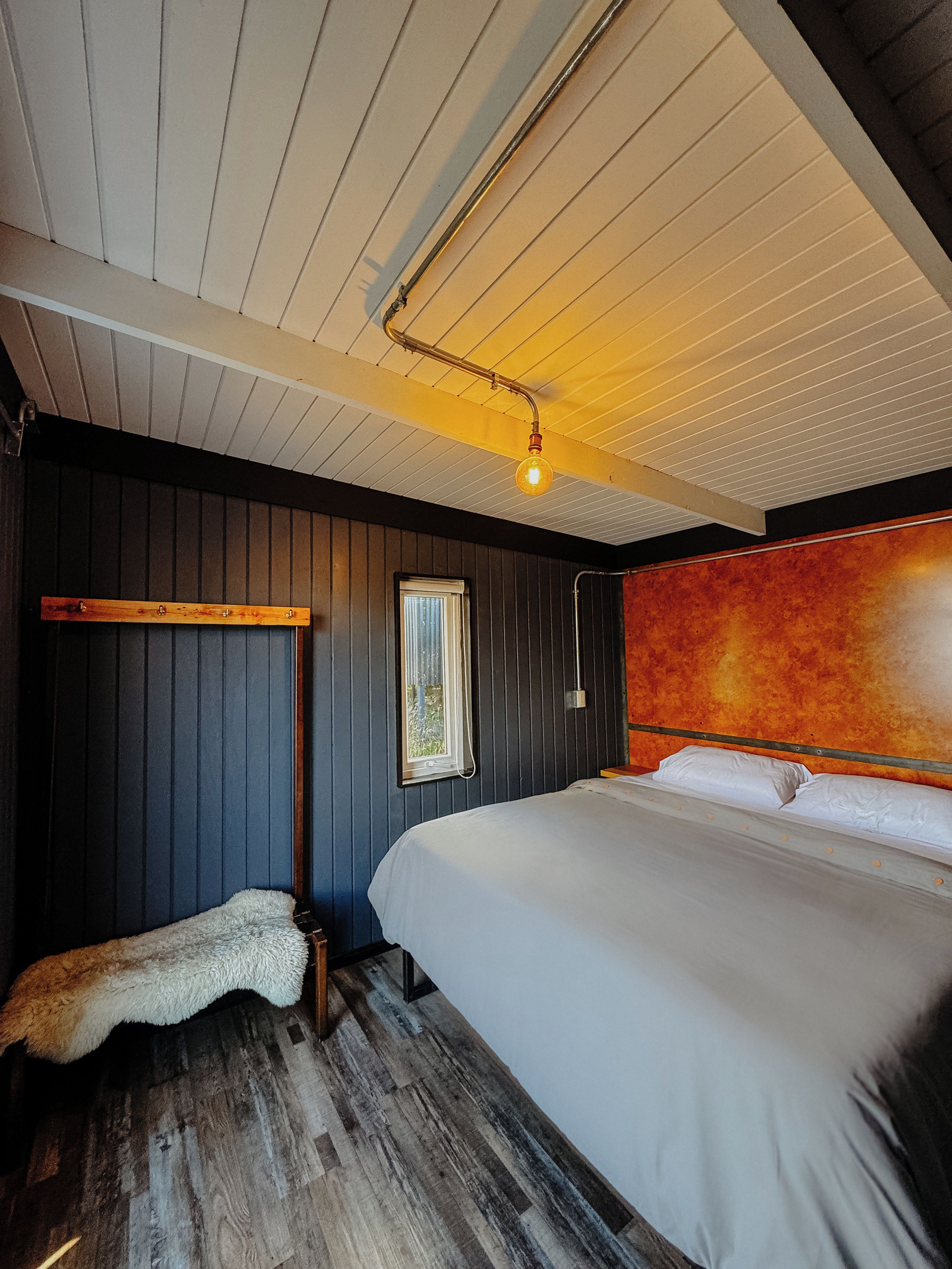 A bedroom with a wooden floor, a bed with white bedding, a hanging light fixture, and a window with blinds.