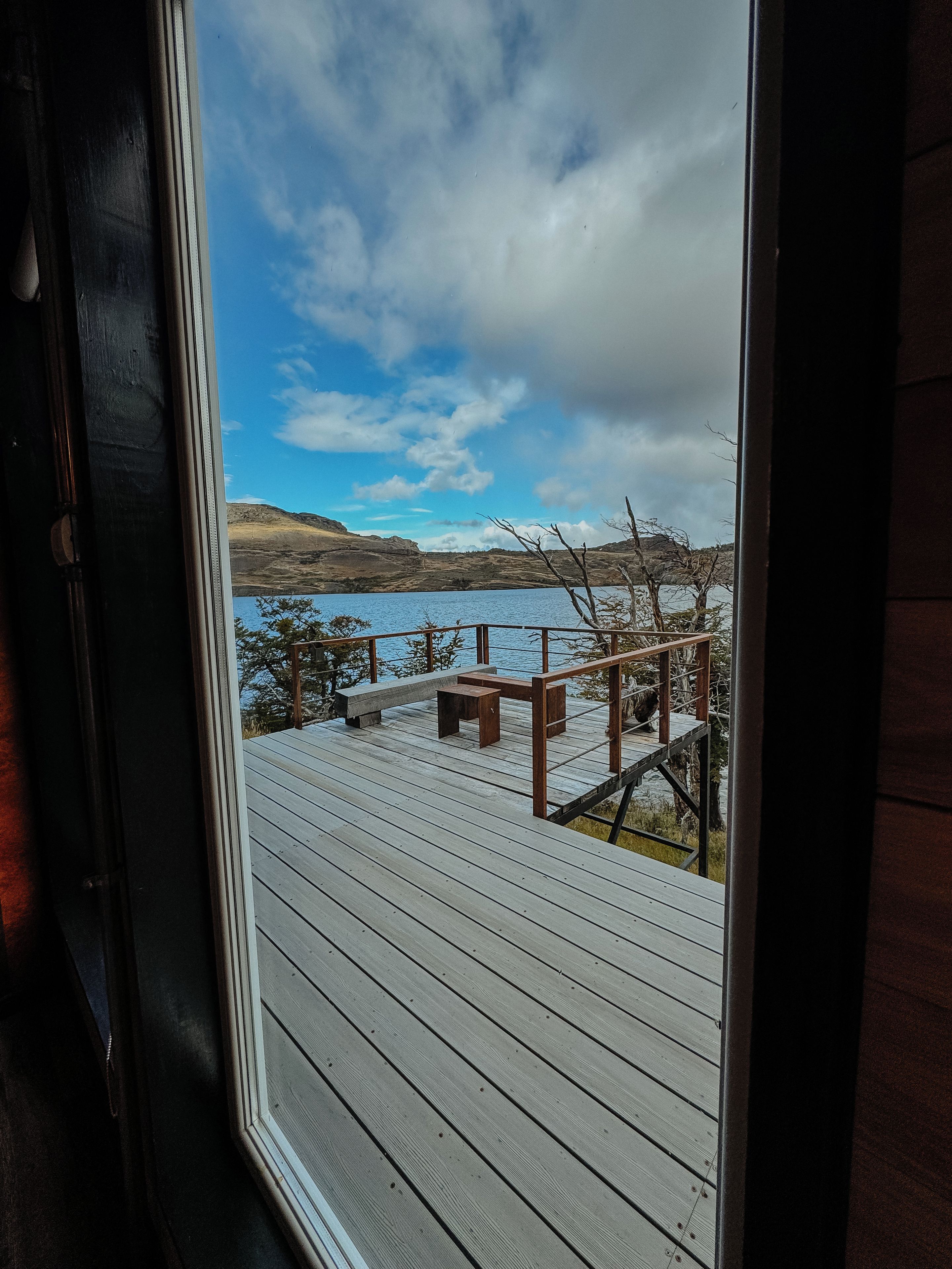 A wooden deck with a bench and a table, overlooking a body of water and hills.