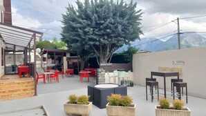 Een terras met tafels en stoelen, een grote boom en uitzicht op een berg.