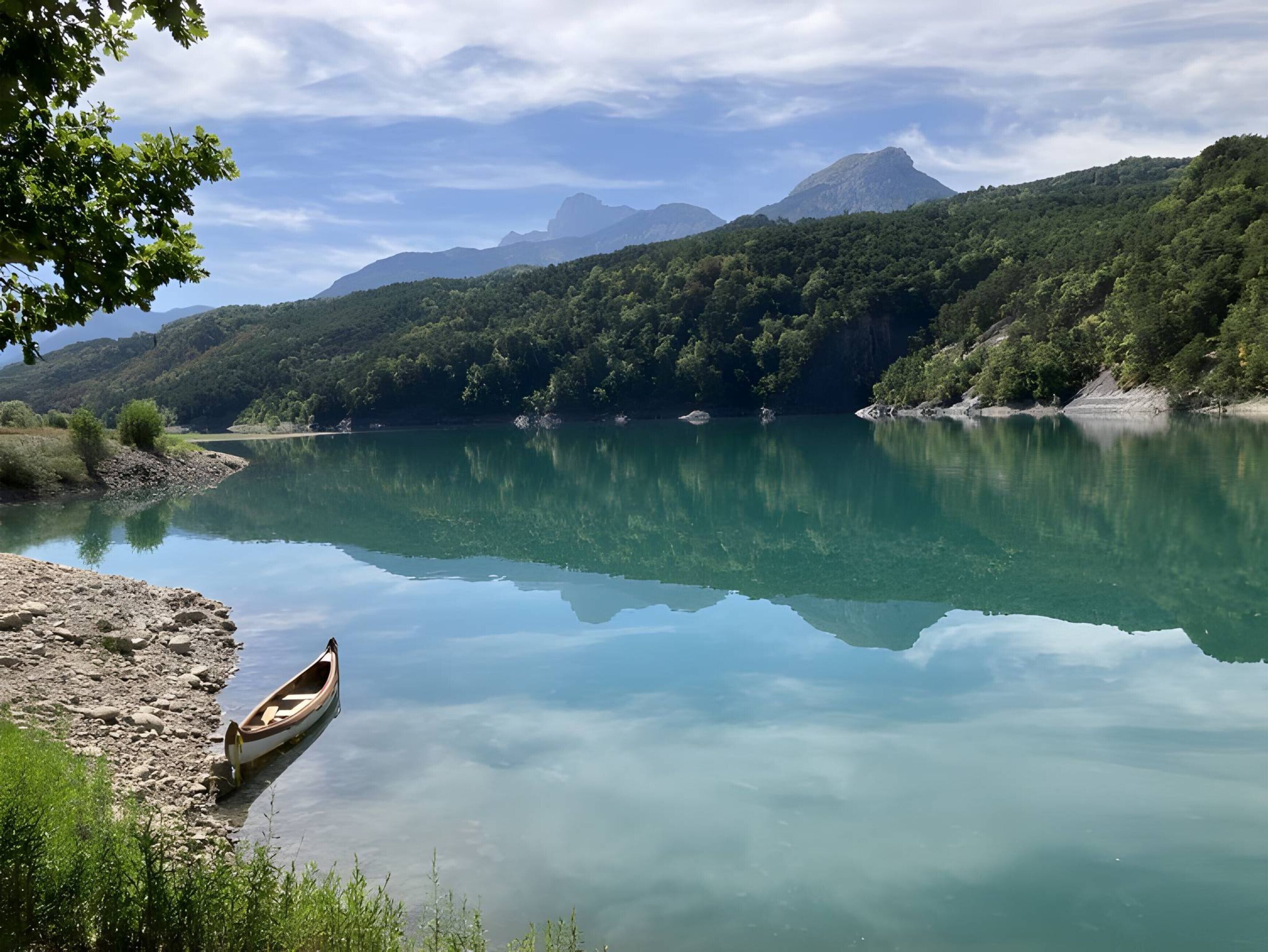 A calm lake with a wooden boat on the shore, surrounded by lush green hills and mountains under a partly cloudy sky.