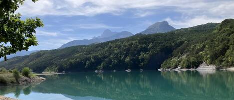 A calm lake with a wooden boat on the shore, surrounded by lush green hills and mountains under a partly cloudy sky.