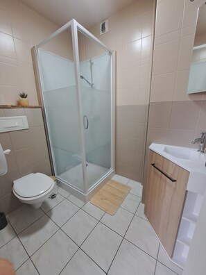 Une salle de bain comprenant des toilettes, une douche fermée par une paroi de verre, un lavabo et une petite étagère sur laquelle est posée une plante.
