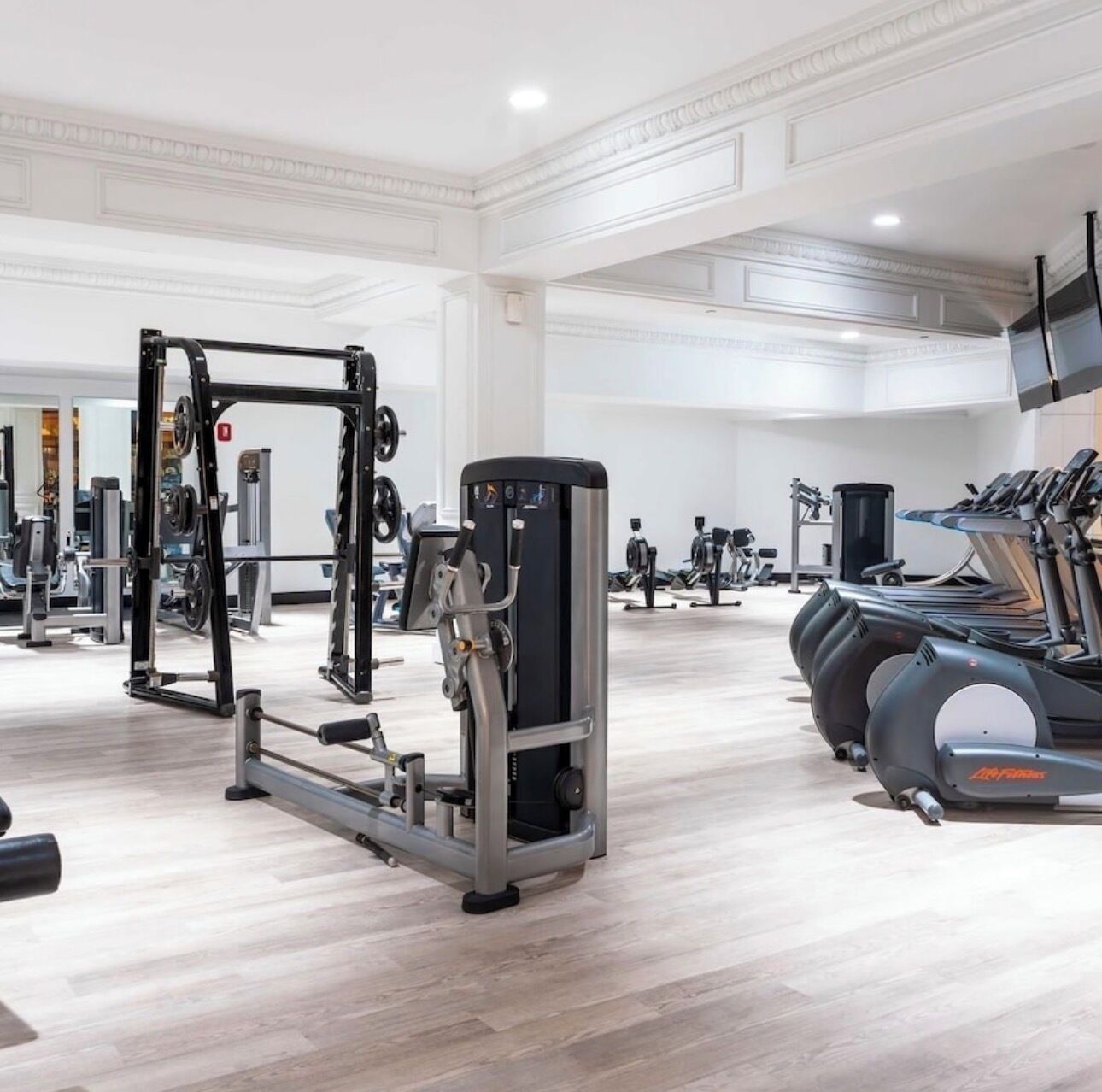 Un gimnasio con varios equipos de ejercicio, incluyendo una estructura para sentadillas, una máquina de press de pecho y cintas de correr.