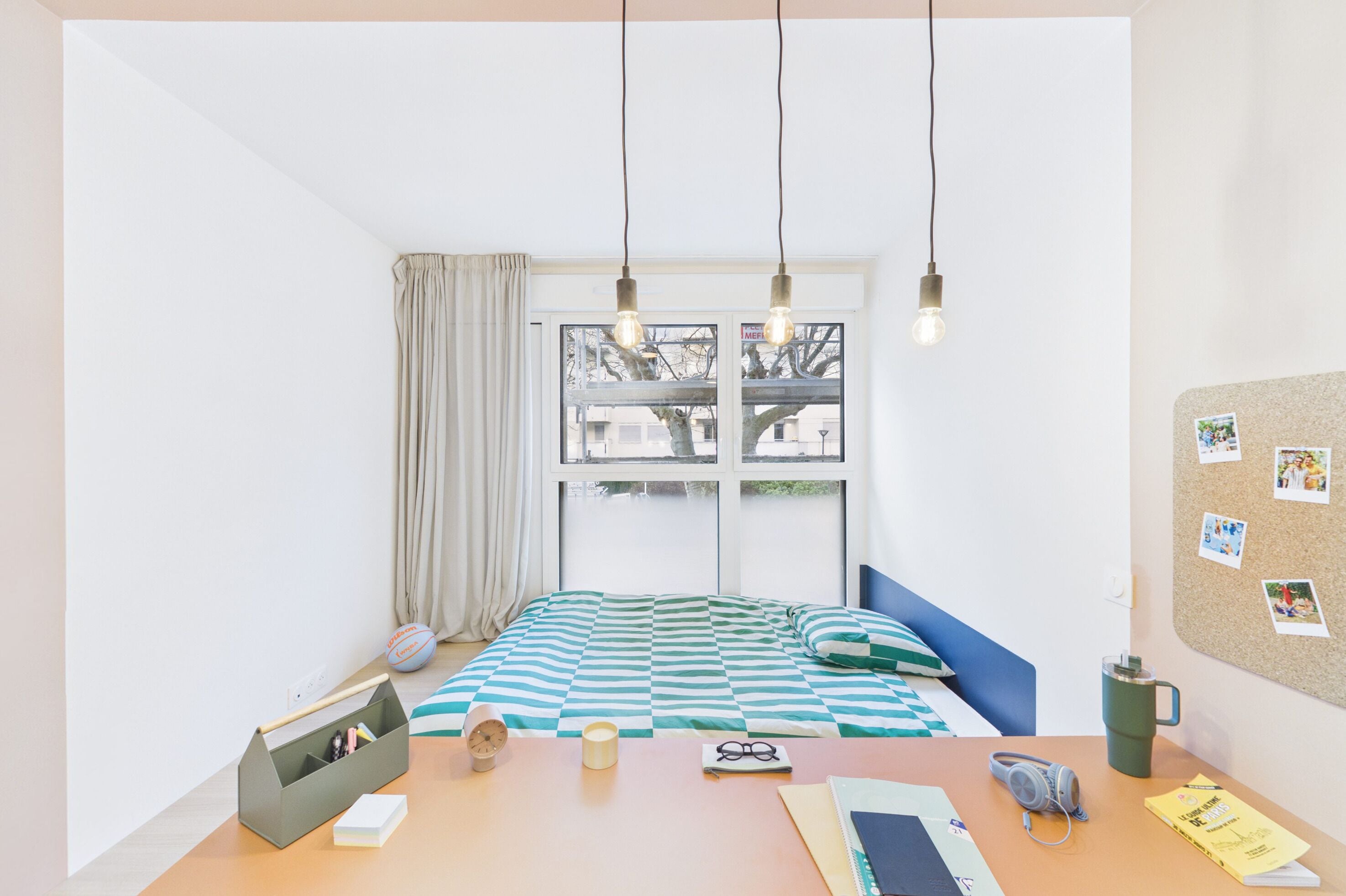 Ein Schlafzimmer mit Bett, Schreibtisch, Fenster und einer Korkwand mit Fotos.
