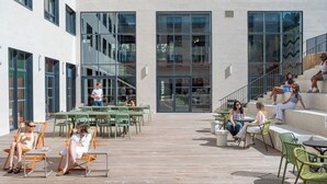 An outdoor seating area with tables and chairs, a wooden deck, and a building with large windows and a staircase.