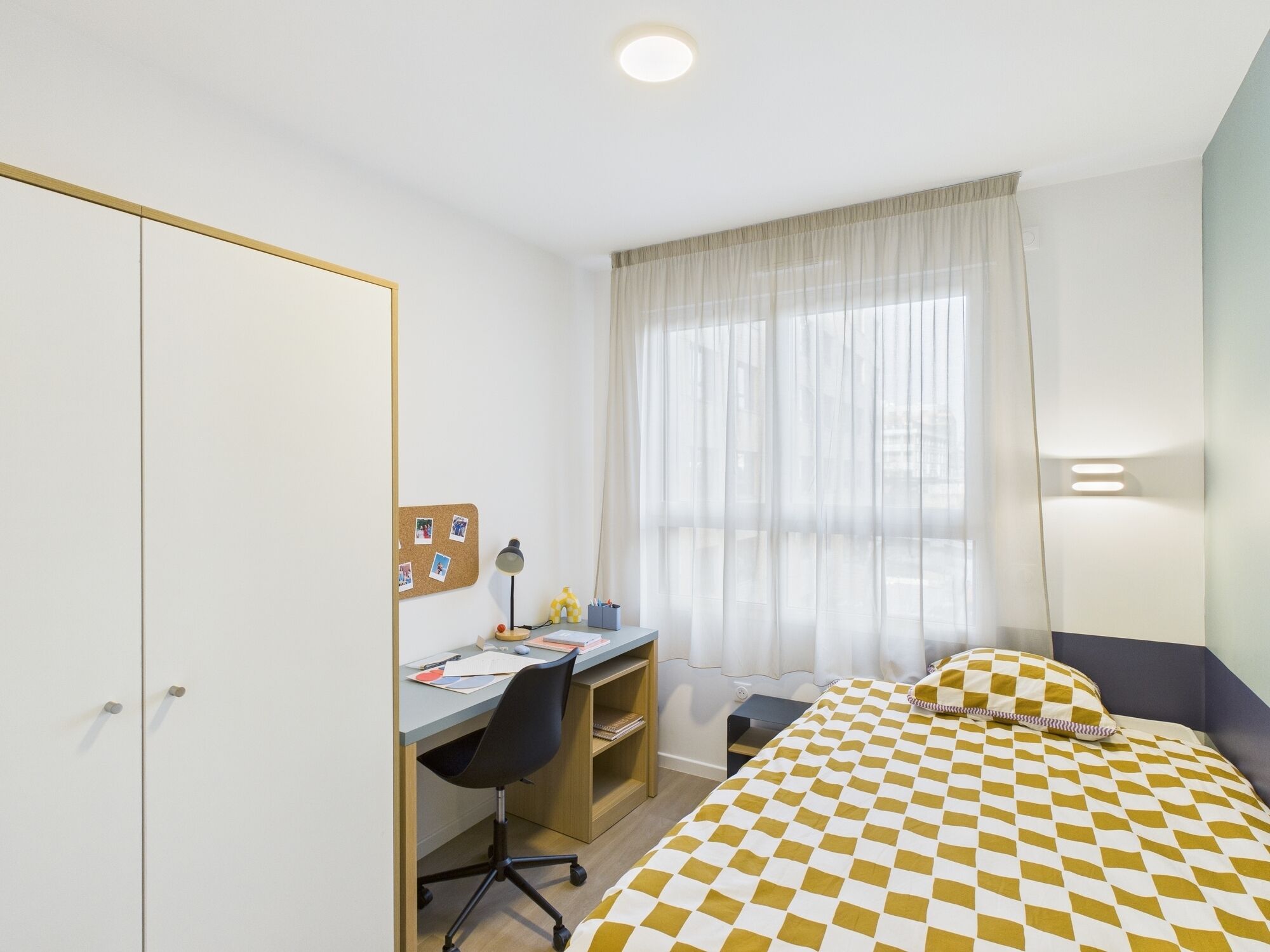 A bedroom with a bed, a desk, a chair, a wardrobe, and a window with curtains.