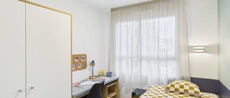 Un dormitorio con cama, escritorio, silla, armario y ventana con cortinas.