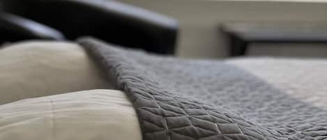 Una cama con un edredón acolchado y almohadas, una ventana con vistas a un edificio y follaje verde.