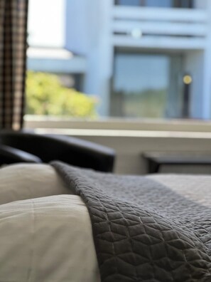 A bed with a quilted blanket and pillows, a window with a view of a building and greenery. - Hotel Lakolk (Rømø)