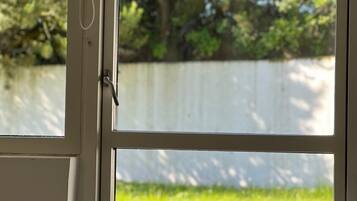 A window with a view of a white fence and trees, a bed with a striped bedspread, and a chain hanging from the window.