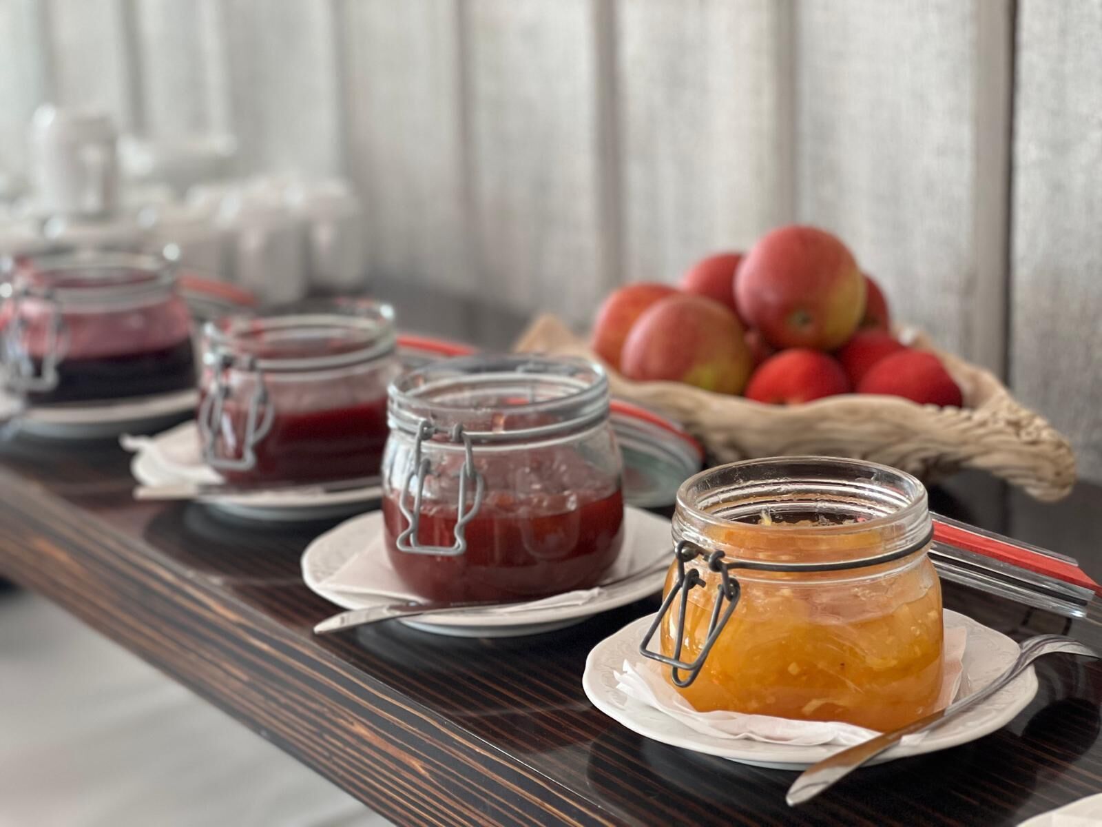 A table with three jars of preserves, a basket of apples, and a white wall in the background.