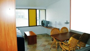 A room with a dining table, chairs, a television, and a window with blinds. - Hotel Lakolk (Rømø)