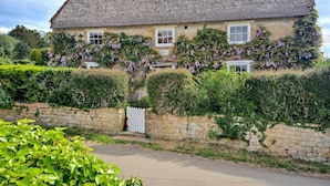 Ein traditionelles Steinhaus mit einem steilen, strohgedeckten Dach, efeuüberwucherten Mauern und einem weißen Ziergittertor.