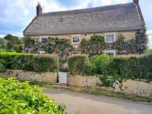 Ein traditionelles Steinhaus mit einem steilen, strohgedeckten Dach, efeuüberwucherten Mauern und einem weißen Ziergittertor.