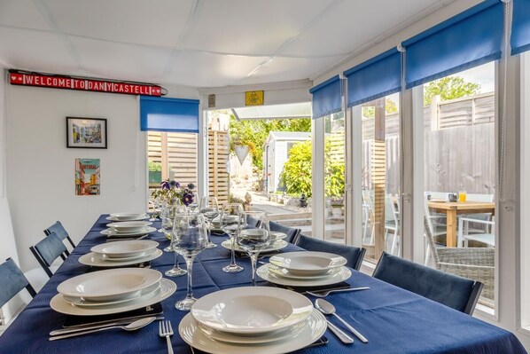 A dining area with a table set for six, blue tablecloth, and a view of the garden through large windows.