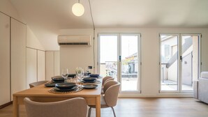 Ein moderner Essbereich mit einem Holztisch, gedeckt für sechs Personen, einem großen Fenster und Blick auf ein Gebäude mit Balkonen.