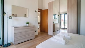 Una camera da letto moderna con un comò in legno, un letto con lenzuola bianche, uno specchio grande e vista sul balcone.