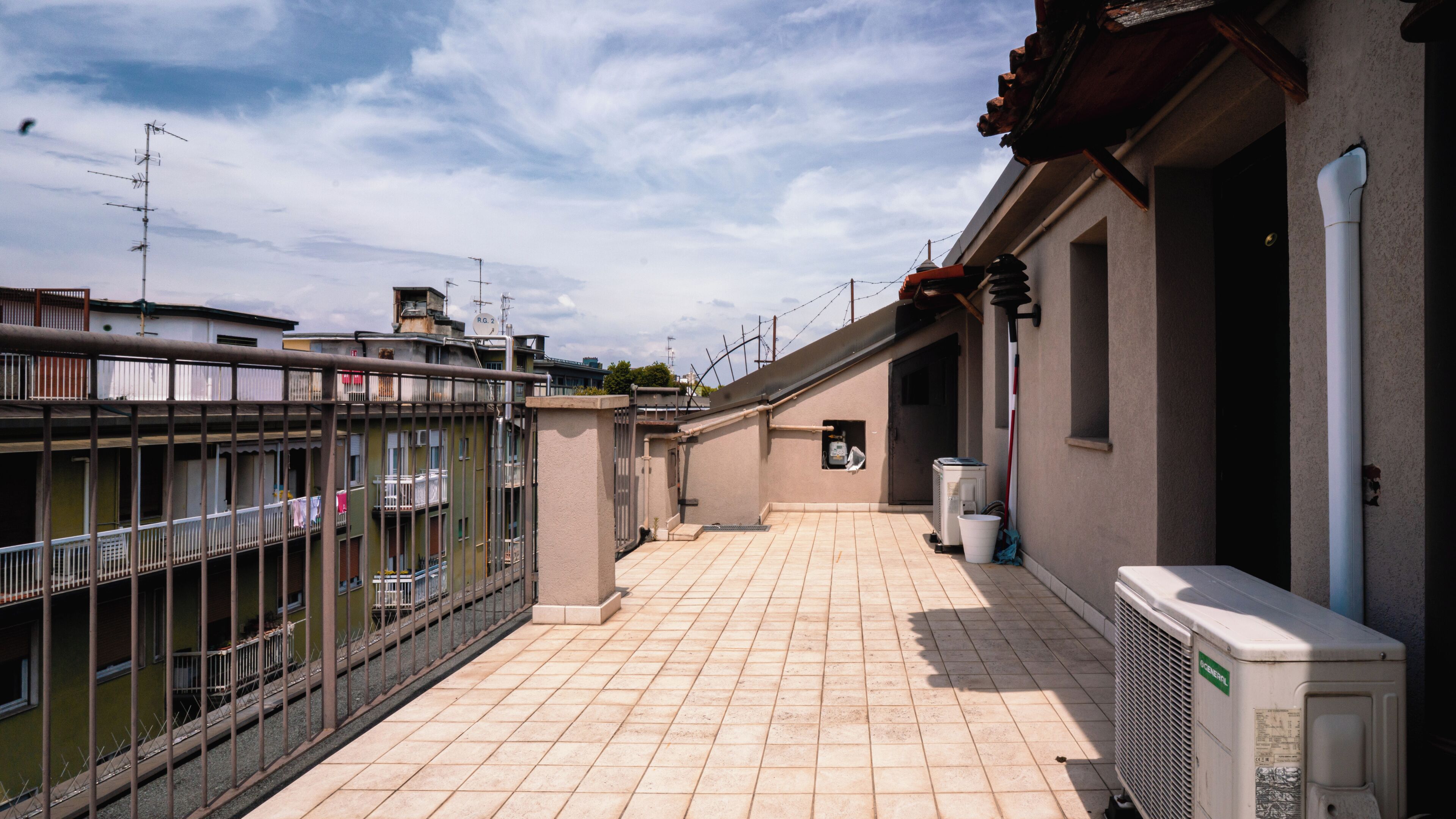 A rooftop terrace with a tiled floor, a white air conditioning unit, and a metal fence.