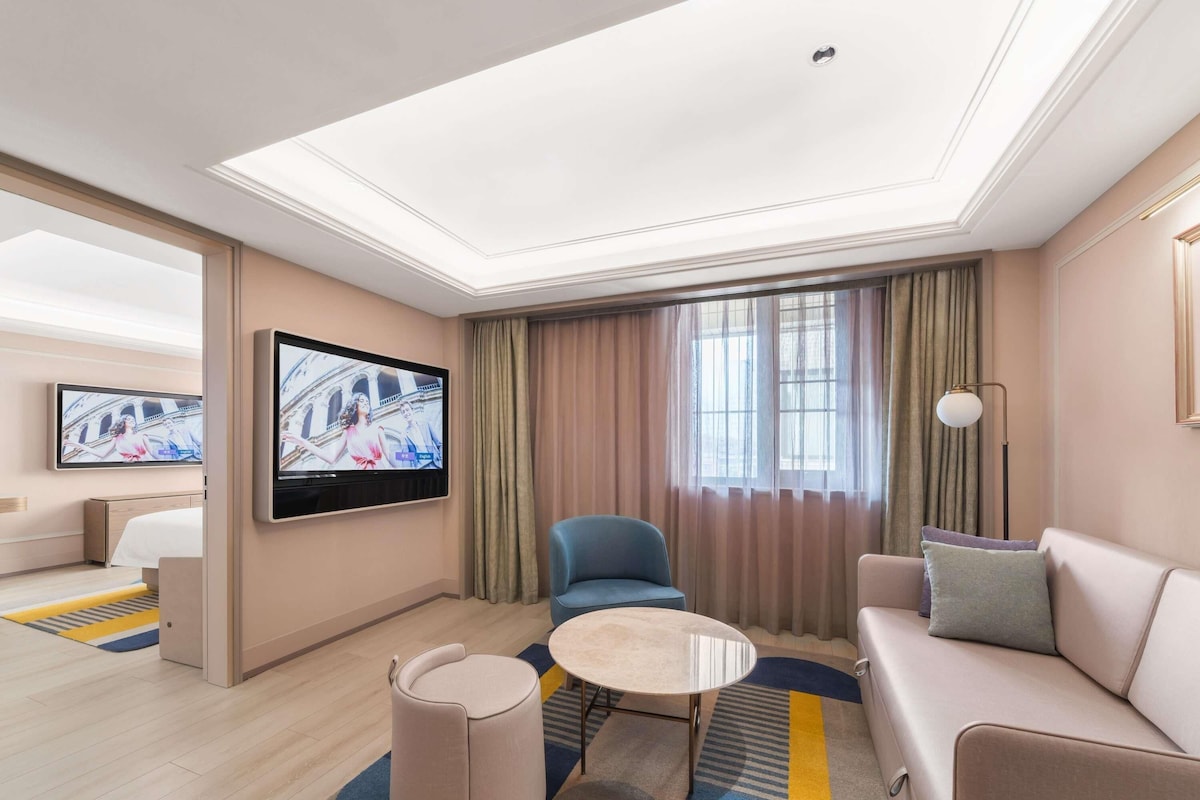 Et moderne hotelværelse med et fladskærms-tv på væggen, en sofa, en blå lænestol, et lille rundt bord og en gulvtæppe med et mønster.