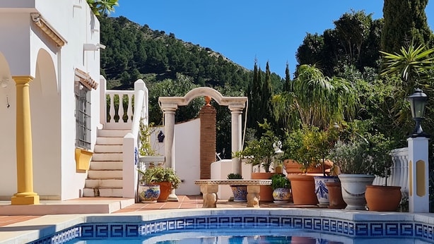 Une piscine ornée d’un motif géométrique en mosaïque, un bâtiment blanc agrémenté d’accents jaunes et, en arrière-plan, un paysage montagneux.