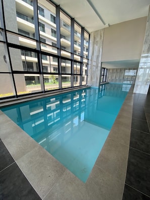Ein Schwimmbad mit Fliesenboden, großen Fenstern und Blick auf ein mehrgeschossiges Gebäude.