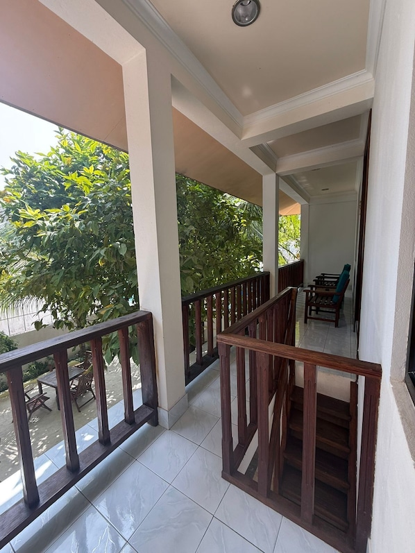 A covered balcony with a wooden railing, a tiled floor, and a view of greenery outside.