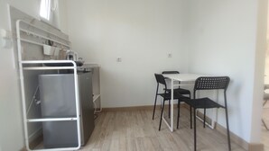 Eine kleine Küchenzeile mit weißer Arbeitsplatte, einem Kühlschrank und einem Spülbecken. Zwei schwarze Stühle und ein weißer Tisch sind ebenfalls vorhanden.