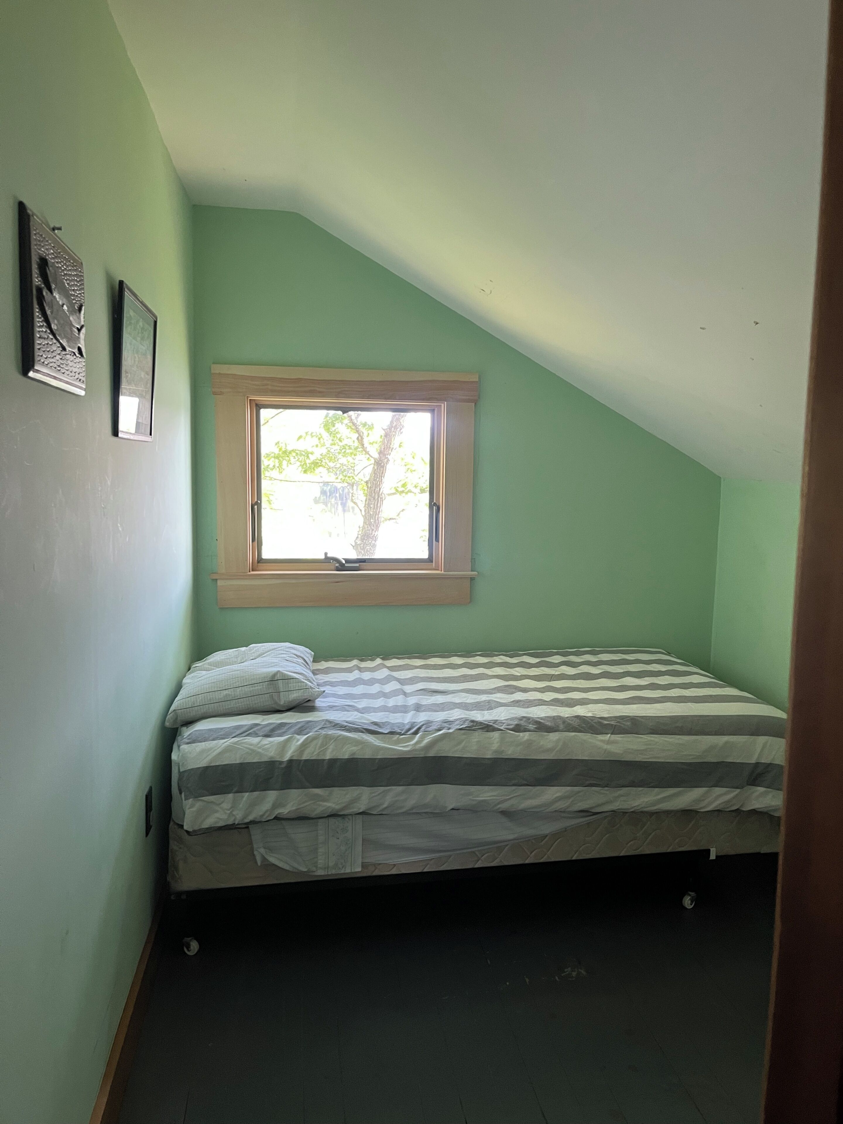 Una piccola camera con un letto singolo, soffitto spiovente, una finestra con cornice in legno e una porta.