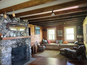 L’intérieur d’un chalet accueillant, avec une cheminée en pierre, des poutres en bois, un mur de pierre, un canapé et une fenêtre ornée d’œuvres d’art.