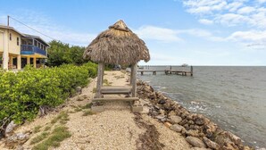 A beach with a thatched structure, a pier extending into the water, and a building with a balcony. - The Peninsular House – Private Waterfront Paradise (Marathon)
