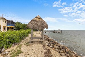 A beach with a thatched structure, a pier extending into the water, and a building with a balcony. - The Peninsular House – Private Waterfront Paradise (Marathon)