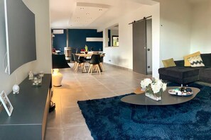 Ein modernes Wohnzimmer mit einem dunkelblauen Teppich, einem Couchtisch mit Blumen, einer Couch mit Kissen und einem Essbereich im Hintergrund.