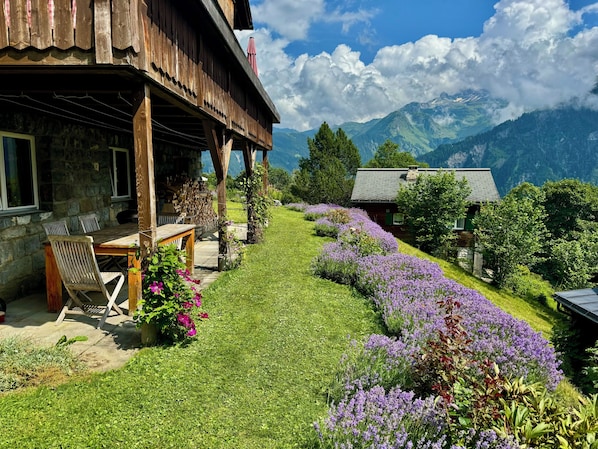 A wooden building with a stone exterior, a covered outdoor seating area, and a garden with purple flowers.
