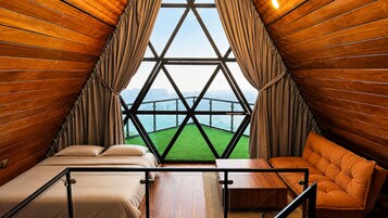 Una habitación con cama, mesa y sofá, con vista a un paisaje verde a través de una ventana geométrica.