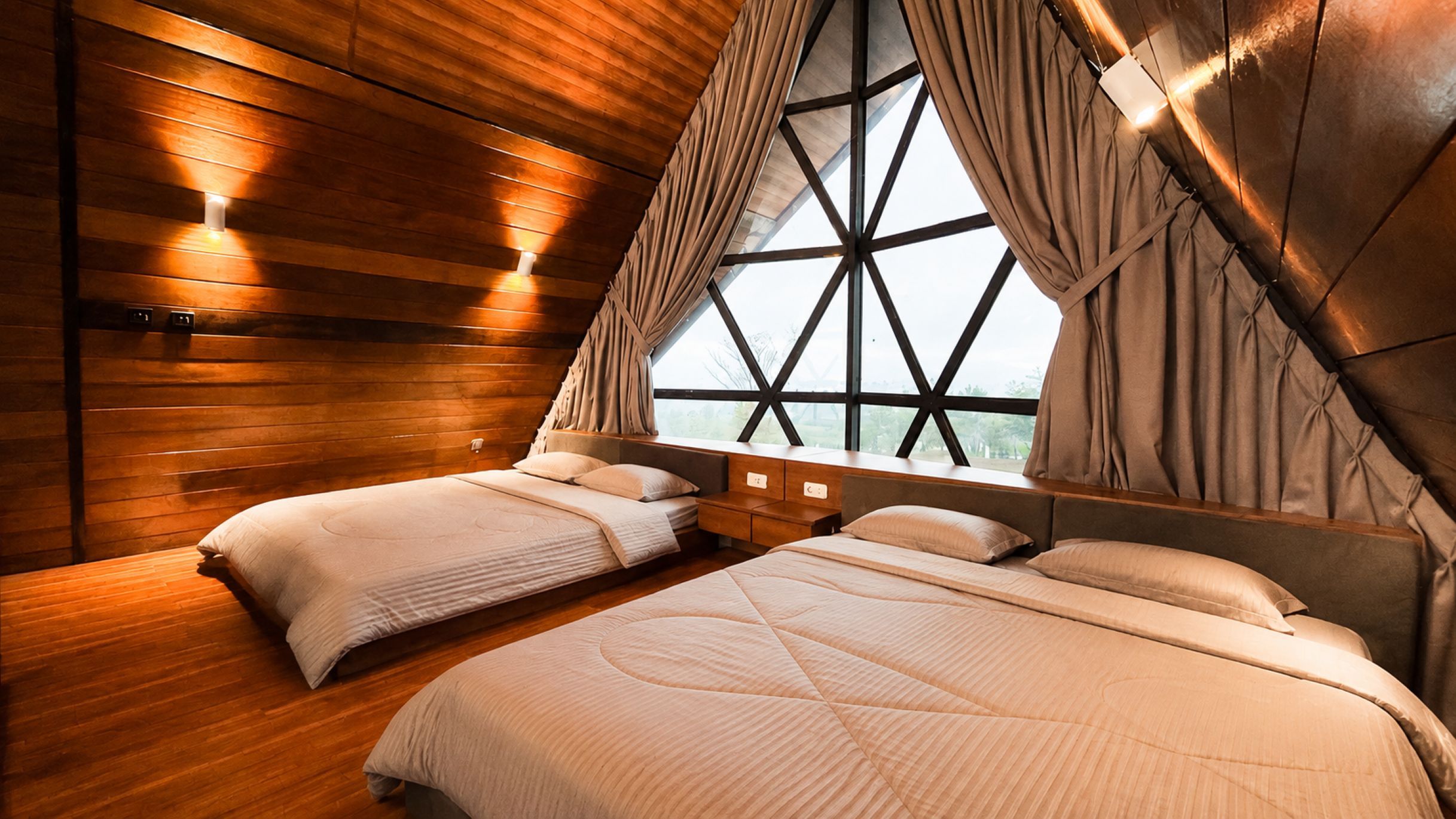 A bedroom with two beds, wooden walls, a large window with curtains, and wall-mounted lights.