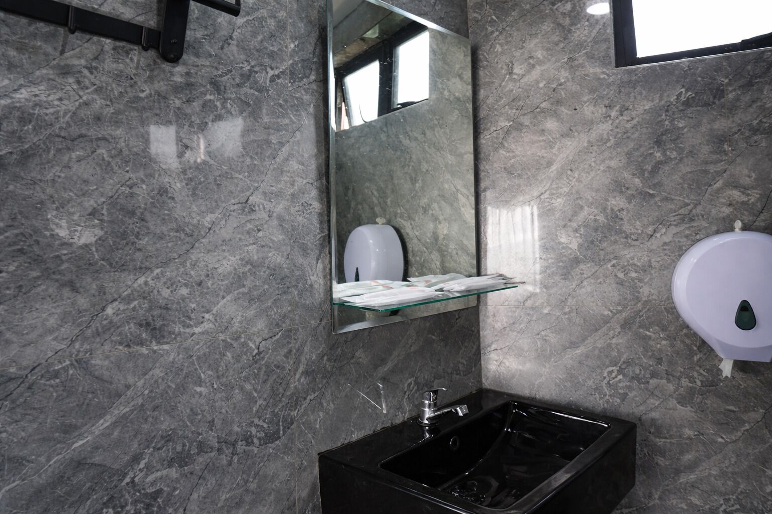 Un baño moderno con pared de mármol, repisa de vidrio con artículos de aseo, lavamanos negro y secador de manos.