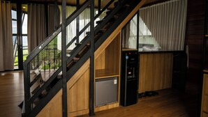 Una escalera con barandilla de vidrio, un refrigerador compacto y un armario de madera en una habitación con ventanas amplias y piso de madera.