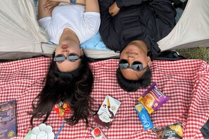 Dos personas descansando en una hamaca sobre una manta a cuadros rojos y blancos, con aperitivos y un libro.