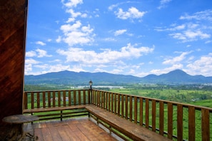 A wooden balcony with a view of green fields and mountains under a blue sky.