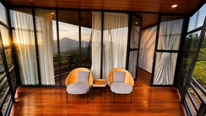 Una habitación moderna con piso de madera, dos sillas con cojines, una mesita pequeña y ventanas grandes con cortinas.