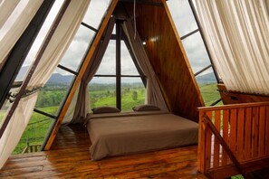 Ein Zimmer mit einem Bett, Holzboden und großen Fenstern mit Blick auf eine grüne Landschaft.