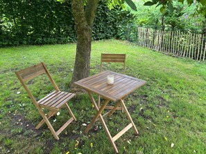 Ein Holztisch und zwei Stühle stehen auf einer grünen Rasenfläche, im Hintergrund ein Baum und ein Holzzaun.