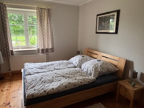 Ein Schlafzimmer mit einem hölzernen Bett, einem Fenster mit Vorhängen, einem Heizkörper, einem Nachttisch mit Lampe und einem gerahmten Bild an der Wand.