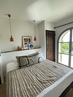 A bedroom with a large bed, two pendant lights, a wooden door, and a view through a window. - Villa CAMILLE proche plage de sable (Ersa)